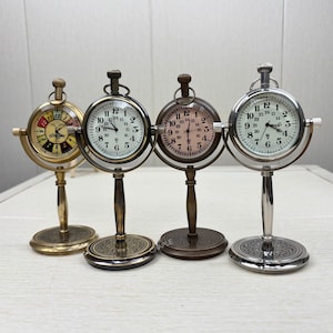 May include: Three antique-style pocket watches on decorative stands. The watches have different colored faces: white, pink, and a colorful roulette wheel design. The stands are made of metal and have a detailed design.