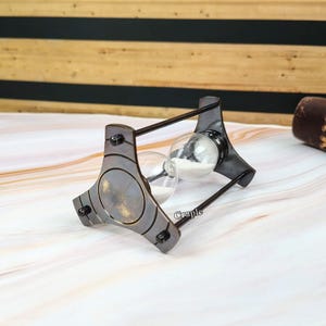 May include: An antique-style hourglass with a dark metal frame and clear glass bulbs. The hourglass is supported by a triangular base with decorative rivets. The sand inside is white. The word "Craple" is printed on the side.