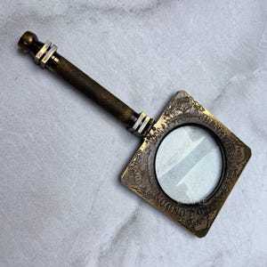 Op de afbeelding: Antieke messing vergrootglas met een ruitvormig frame en een getextureerde handgreep. Het frame is gedetailleerd met reliëfpatronen en tekst. De vergrootlens is rond en helder.