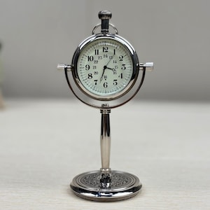 Pode incluir: Um relógio de mesa prateado com um mostrador redondo e numerais romanos pretos. O relógio tem um suporte com uma base decorativa.