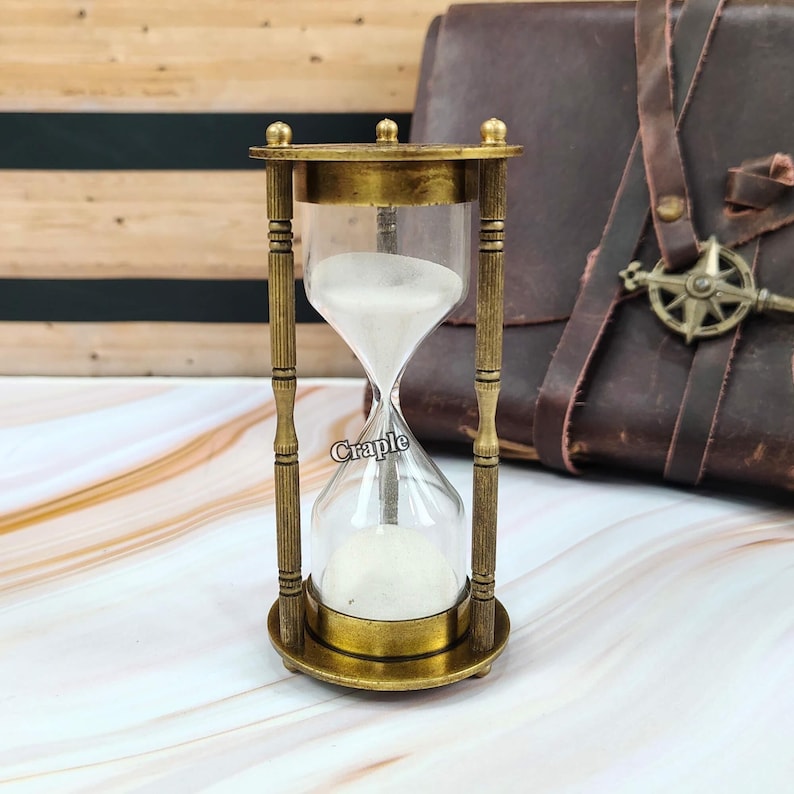 May include: An antique-style hourglass with a brass frame and clear glass bulbs. The hourglass contains white sand and has the word "Craple" printed on the glass. The design features decorative vertical bars and a round base.