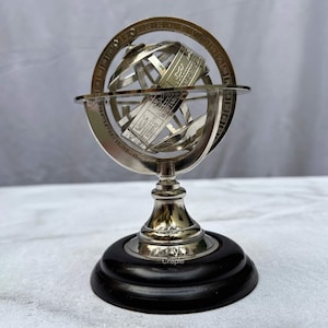 May include: A silver-colored armillary sphere with a black wooden base. The sphere features intricate metal bands and a central section with zodiac signs. The base is round and has a polished finish. The sphere is a decorative item.