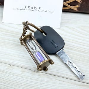 May include: A vintage-style hourglass keychain with a brass finish, filled with purple sand. The keychain is attached to a car key. The card in the background reads "CRAPLE Handcrafted Antique & Nautical Decor."