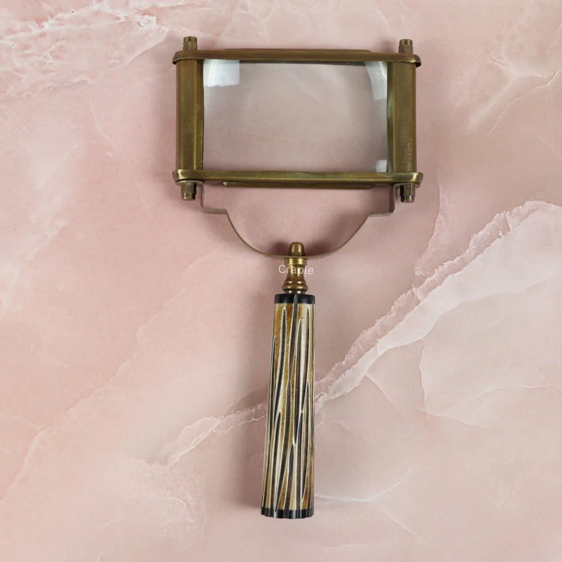 May include: A vintage-style magnifying glass with a rectangular lens set in a brass frame. The handle features a striped pattern in cream, black, and gold. The magnifying glass is designed for reading or detailed inspection.
