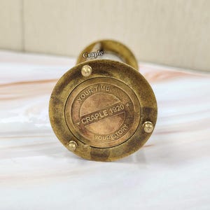 May include: A vintage brass hourglass with the text "YOUR TIME YOUR STORY CRAPLE 1920" on the base. The hourglass has a classic design with a circular base and top, and a narrow center.