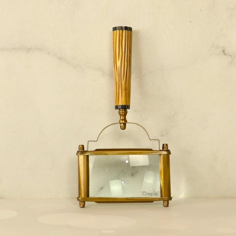 May include: A vintage brass magnifying glass with a rectangular lens and a wooden handle. The handle is fluted and has a dark accent at the top. The word "Craple" is printed on the glass.