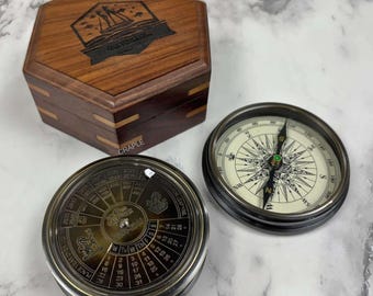 May include: A wooden hexagonal box with a ship emblem, and two compasses. One compass has a white face with black markings, and the other is a calendar compass with a rotating dial. The box has the word "CRAPLE" on it.