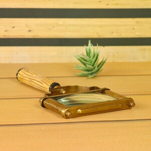 May include: A vintage-style brass magnifying glass with a wooden handle. The magnifying glass has a rectangular frame and a clear lens. The handle is light brown with a textured design. The word "Crapie" is engraved on the frame.
