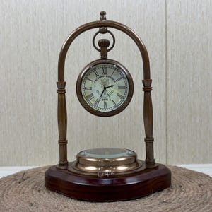 Op de afbeelding: Een klok in vintage-stijl met een ronde wijzerplaat, Romeinse cijfers en de tekst "VICTORIA LONDON 1876". De klok is opgehangen in een bronskleurig gebogen frame, gemonteerd op een donkere houten basis. Een vergrootglas bevindt zich aan de basis.