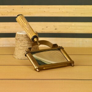 May include: A vintage brass magnifying glass with a rectangular lens and a wooden handle. The handle is light brown with a textured design. The magnifying glass is resting on a wooden surface next to a small, twine-wrapped object.