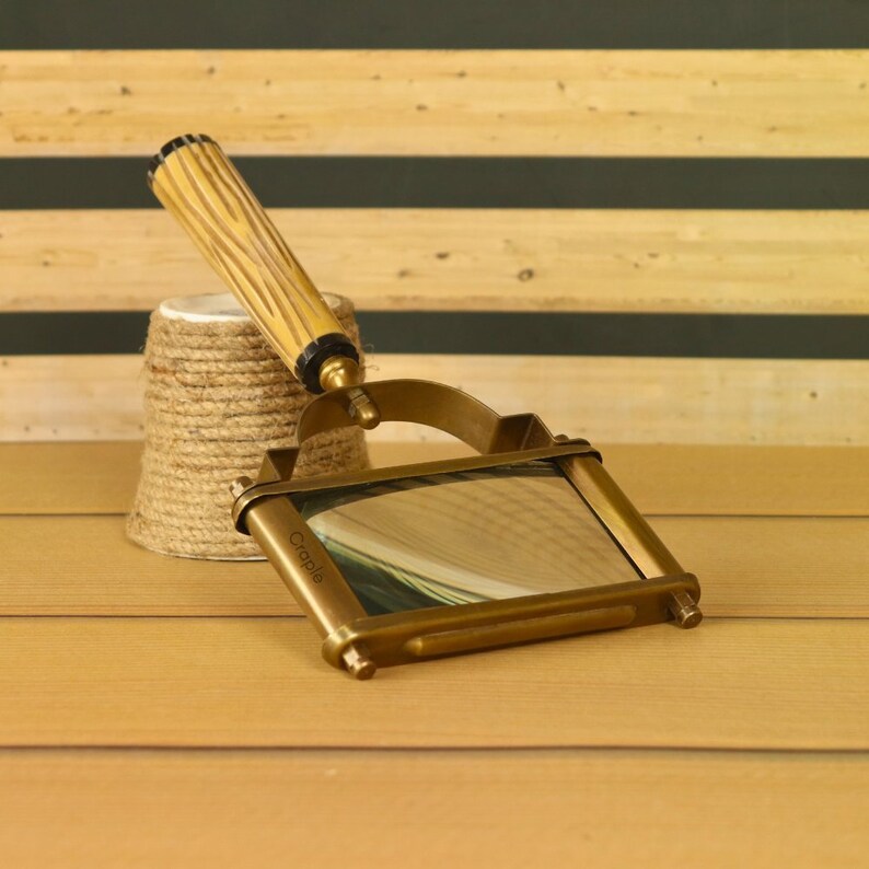 May include: A vintage brass magnifying glass with a rectangular lens and a wooden handle. The handle is light brown with a textured design. The magnifying glass is resting on a wooden surface next to a small, twine-wrapped object.