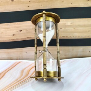 May include: An antique-style hourglass with a brass-colored frame and clear glass chambers. The hourglass is filled with white sand. The word "Craple" is printed on the glass. The hourglass is a decorative item.