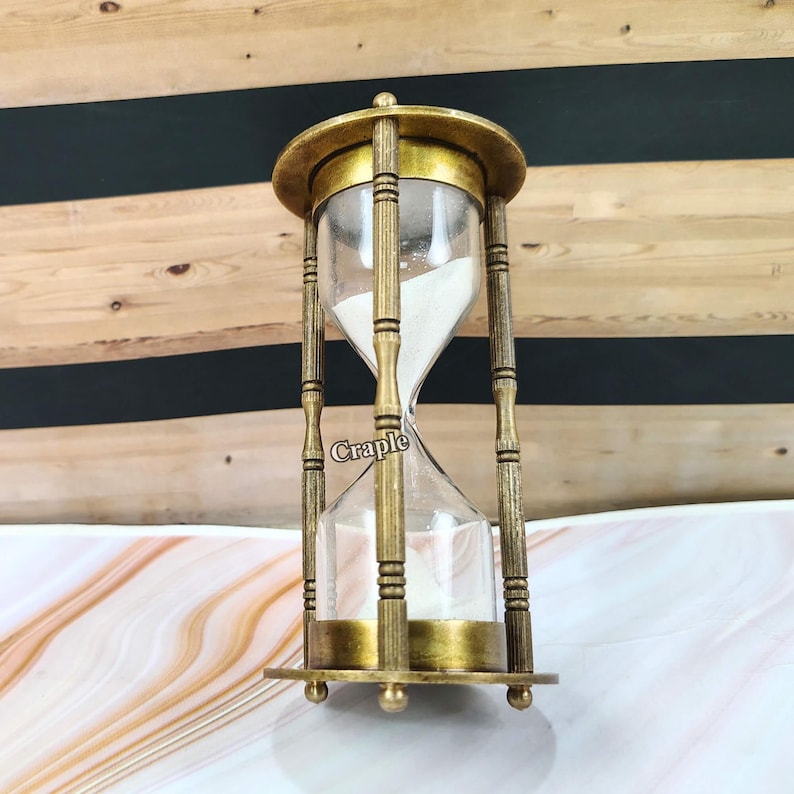 May include: An antique-style hourglass with a brass-colored frame and clear glass chambers. The hourglass is filled with white sand. The word "Craple" is printed on the glass. The hourglass is a decorative item.