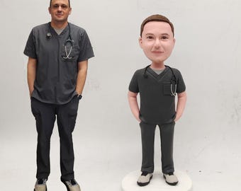 May include: A person wearing a dark gray V-neck scrub top and matching pants with a stethoscope around their neck, standing next to a custom bobblehead figurine that replicates their appearance, clothing, and stethoscope in a miniature form.
