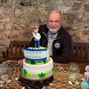 May include: A two-tier white frosted cake decorated with green foliage and grey stones, topped with a figurine of a person holding a fish. A banner on the cake reads Fishing You A HAPPY RETIREMENT. The cake sits on a wooden stand on a table.