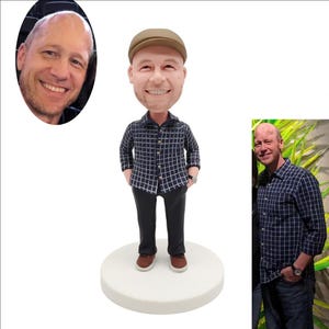 May include: A custom bobblehead figurine depicting a smiling person wearing a tan flat cap, a dark blue button-down shirt with a white grid pattern, dark trousers, and brown shoes, standing on a circular white base, accompanied by two reference photos.