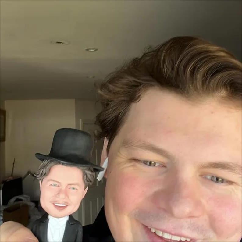 May include: A small figurine featuring a person with brown hair wearing a black top hat, a white collared shirt, and a black suit jacket. The figurine has a smiling facial expression and is held in front of a person with brown hair.