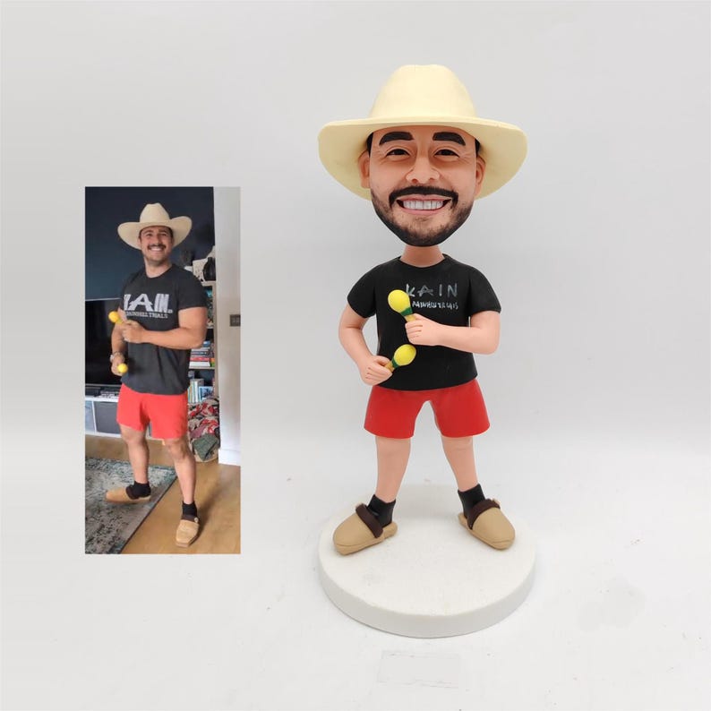 May include: A custom bobblehead figurine depicting a person wearing a light-colored cowboy hat, a black t-shirt with the text KAIN, red shorts, and tan slippers. The figure holds two small yellow maracas and stands on a circular white base.