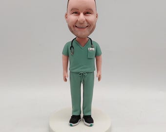 May include: A bobblehead figurine of a smiling person in green medical scrubs, a stethoscope, and black sneakers. The figurine is standing on a white circular base. The head is oversized.