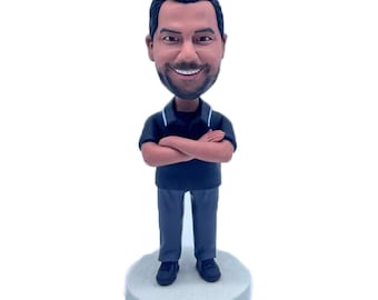 May include: A custom bobblehead figurine of a person with a black polo shirt, grey trousers, and a smiling face. The figurine is standing with arms crossed and is on a white base.
