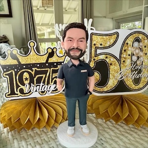 May include: A custom bobblehead figurine of a bearded person wearing a dark polo shirt and gray trousers, standing on a white circular base. Behind the figure are gold glittery 50th birthday signs with the text 1975 Vintage and Happy Birthday.