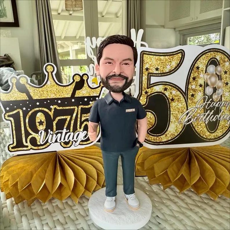 May include: A custom bobblehead figurine of a bearded person wearing a dark polo shirt and gray trousers, standing on a white circular base. Behind the figure are gold glittery 50th birthday signs with the text 1975 Vintage and Happy Birthday.