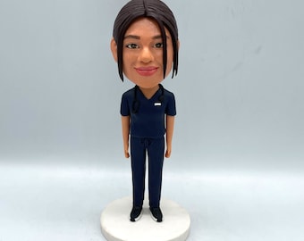 May include: A custom bobblehead figurine of a person wearing a dark blue medical scrubs uniform with a stethoscope around their neck. The figurine has a white base.