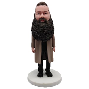 Peut inclure: Figurine d'un homme avec une grande barbe noire, portant un manteau beige, un pantalon noir et des chaussures noires. La figurine repose sur une base circulaire blanche. L'homme a la tête rasée avec un chignon.