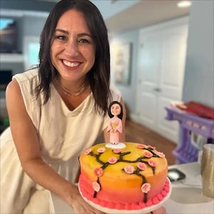 May include: A round cake with an ombre yellow to pink frosting, decorated with dark brown branches and small pink cherry blossom flowers. A small figurine of a person wearing a light pink kimono stands on top of the cake as a decorative topper.