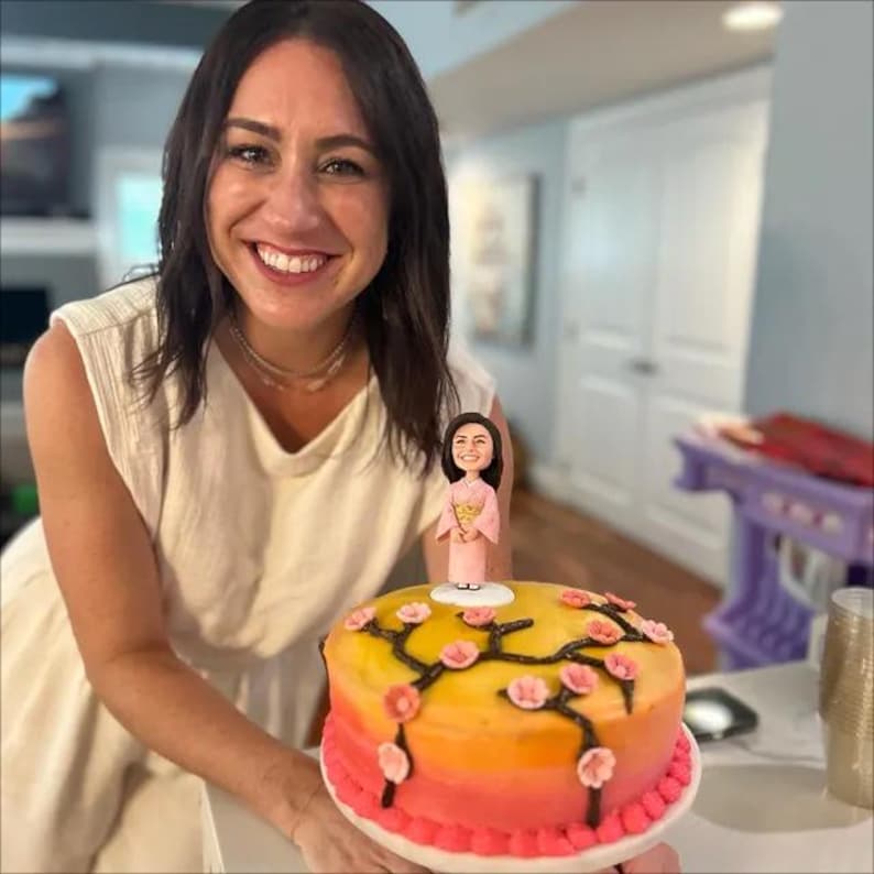 May include: A round cake with an ombre yellow to pink frosting, decorated with dark brown branches and small pink cherry blossom flowers. A small figurine of a person wearing a light pink kimono stands on top of the cake as a decorative topper.
