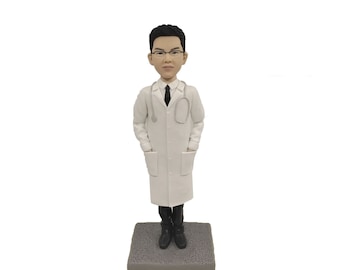 May include: A figurine of a doctor wearing a white lab coat, black tie, and glasses, with a stethoscope around their neck. The doctor is standing on a gray textured base. The figurine has black hair and is wearing black boots.