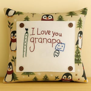 May include: A decorative pillow with a beige background featuring penguins and Christmas trees. A white square is attached with brown buttons, displaying the text "I Love you granpapa" and a drawing.