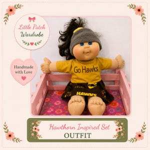May include: A doll with dark hair in a ponytail, wearing a grey beanie, a mustard yellow t-shirt with the text Go Hawks, and a dark brown skirt with the text HAWKS printed on it. The doll is seated in a small pink wooden crate.