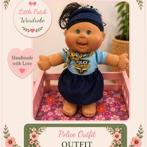 May include: A doll with dark hair and green eyes wearing a light blue shirt with a Police Department badge graphic, a dark blue skirt, and a dark blue headband. The doll has a small patterned neck scarf and is standing in a pink wooden frame.