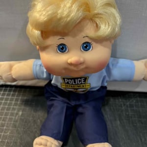May include: A vintage doll with a soft body, blonde curly hair and blue eyes. The doll wears a light blue police shirt with "POLICE DEPARTMENT" and dark blue trousers. The doll's arms and legs are outstretched. A classic toy.