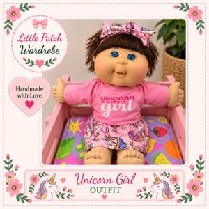 May include: A doll with brown hair and blue eyes wearing a pink long-sleeve shirt with the text UNICORN girl and a matching skirt featuring a unicorn pattern. The doll has a pink unicorn-patterned headband and sits in a small pink wooden crate.