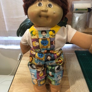 May include: A Cabbage Patch Kid doll wearing a white t-shirt and dungarees with a Star Wars character print. The dungarees are yellow and have a brown strap. The doll has brown curly hair and brown eyes.
