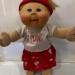 May include: A Cabbage Patch Kid doll wearing a red hat, a white shirt with the words "Go Swans" printed on it, and a red skirt with the Sydney Swans logo.