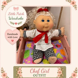 May include: A doll with blonde curly hair and blue eyes wearing a white chef jacket with black and white chequered cuffs, a red necktie, and matching black and white chequered trousers. The doll is seated in a pink wooden crate on a fruit-patterned mat.