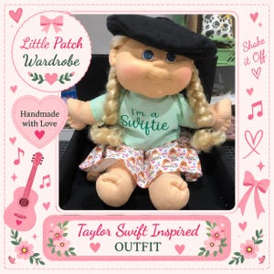 May include: A doll with blonde hair in two braids, wearing a black beret and a light green t-shirt with the text I'm a Swiftie in dark green letters. The doll has a skirt with a colourful pattern and is sitting in a black chair.