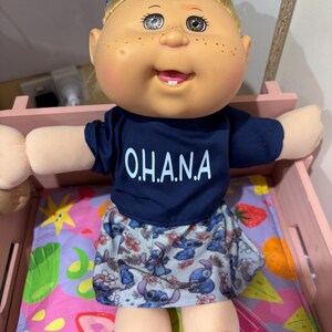 May include: A Cabbage Patch Kid doll wearing a blue shirt with "O.H.A.N.A." written on it, a blue and white skirt with a cartoon character print, and a blue and white headband. The doll is sitting on a pink and purple bed with a fruit print.