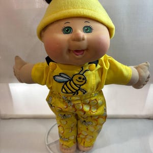 May include: A vintage doll with green eyes and a smiling expression. The doll wears a yellow outfit with a bee design on the front and honeycomb and bee patterns on the trousers. A yellow hat sits atop the doll's brown hair.
