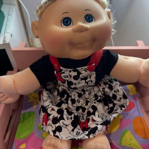 May include: A Cabbage Patch Kid doll with blonde hair wearing a black and white Mickey Mouse print dress with a red bow and a black headband.