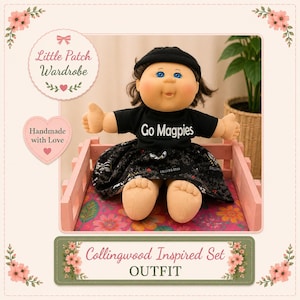 May include: A doll with brown hair and blue eyes wearing a black beanie, a black t-shirt with the white text Go Magpies, and a black skirt featuring a patterned design. The doll is seated in a small pink wooden frame with a floral base.