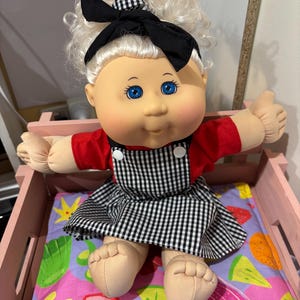 May include: A Cabbage Patch Kid doll with blonde curly hair wearing a red and black checked dress, a black headband with a bow, and blue eyes. The doll is sitting in a pink wooden bed with a colourful fruit-themed bedding.