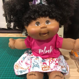 May include: A doll with dark curly hair, wearing a pink t-shirt with the word "pink" and a patterned skirt. The skirt features various graphics and text, including "WHO KNEW" and "LIKE F". The doll has brown eyes and a cheerful expression.