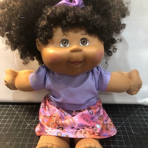 May include: A Cabbage Patch Kid doll with brown skin, brown eyes, and black curly hair. The doll is wearing a purple shirt and a pink and orange patterned skirt.
