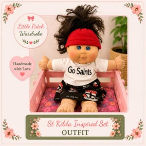 May include: A doll with dark brown hair in a high ponytail, wearing a red headband, a white t-shirt with the text Go Saints, and a black skirt featuring the St. Kilda Football Club logo and text. The doll is seated in a small pink wooden crate.