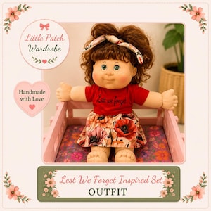 May include: A doll with brown curly hair and a headband, wearing a red t-shirt with the text Lest we forget and a skirt featuring a red poppy floral pattern. The doll is seated in a light pink wooden crate with a floral patterned base.