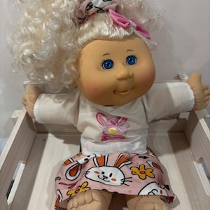 May include: A Cabbage Patch Kid doll with blonde curly hair, blue eyes, and a pink bow. The doll is wearing a white shirt with a pink butterfly design and a pink skirt with a bunny rabbit print.
